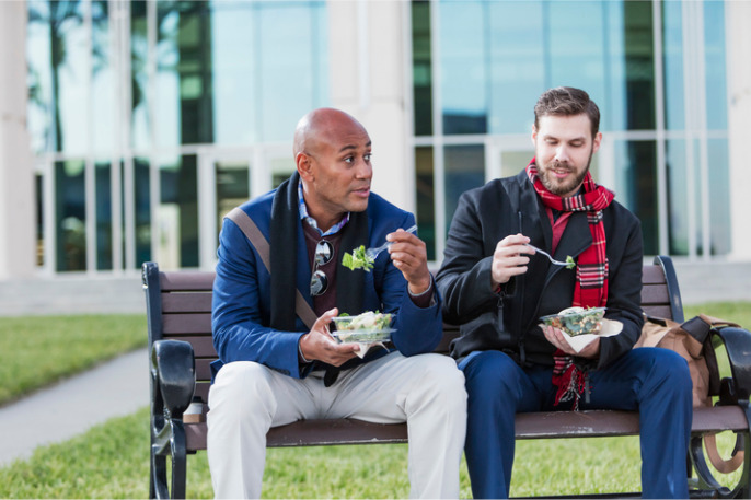 Two men eating lunch on bench outside office building
